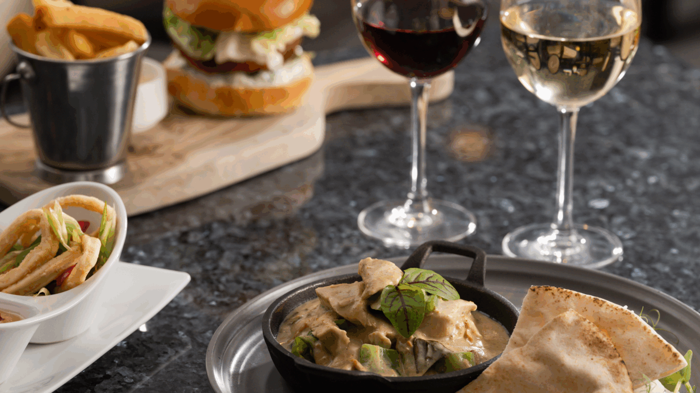 Indulge in hotel dining with a gourmet burger, creamy curry, crispy chips, and fine wines on elegant table setting.