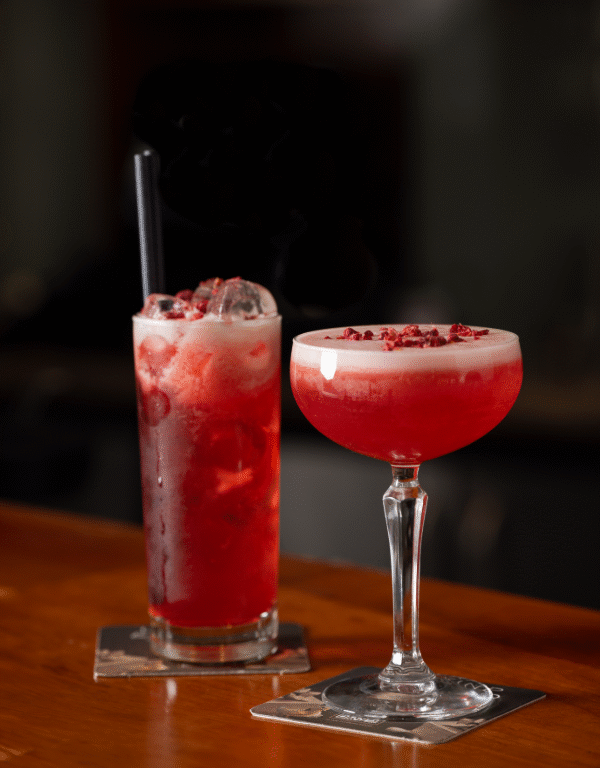 two red cocktails sitting on top of a bar, one in a short cocktail glass and another in a tall glass with a straw