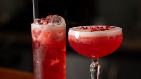 two red cocktails sitting on top of a bar, one in a short cocktail glass and another in a tall glass with a straw