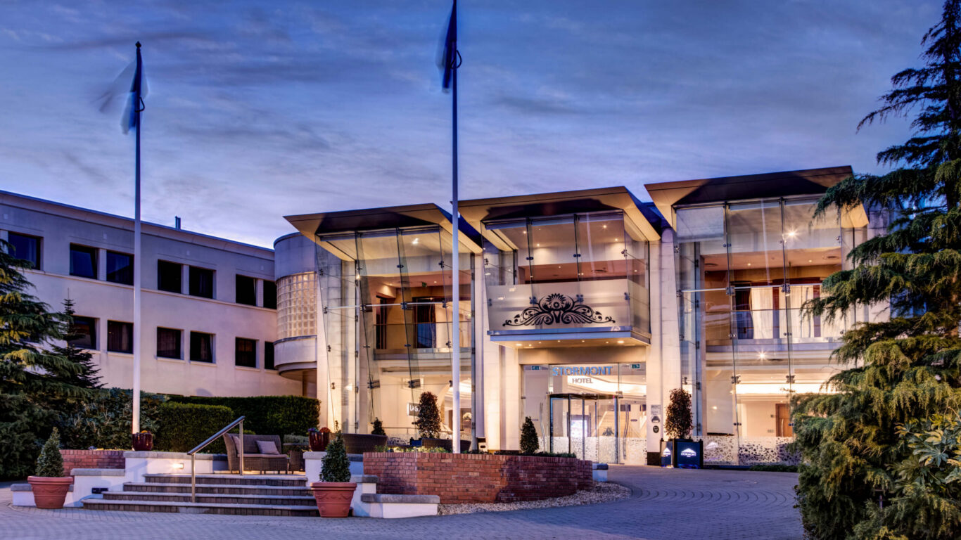 Elegant hotel entrance at dusk with glass façade, welcoming flags, landscaped gardens; ideal for relaxing stays.